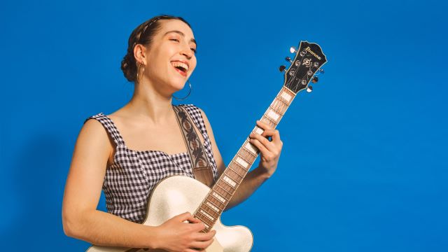 Alisa Amador smiles while singing and playing guitar. She is wearing a gingham dress and is standing if front of a bright blue background.