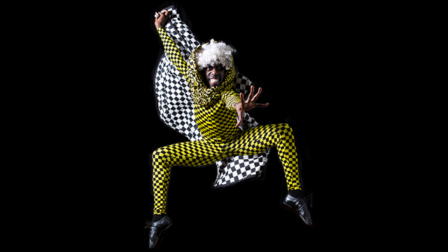 a dancer portraying the mouse king jumps up and reaches one hand out toward the camera. He is wearing a yellow and black checkered suit and is holding a checkered flag above his head. He looks intensely mischievous.