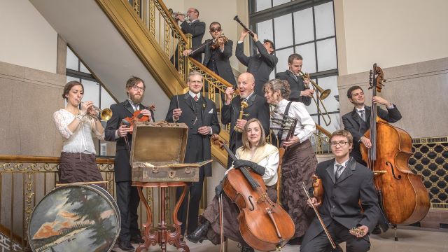 The paragon ragtime orchestra arranged in various poses around one on an old staircase. They are all wearing period clothing and holding their instruments.