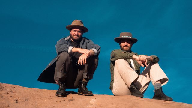 Upward looking picture of two men on top of a dirt hill with the blue sky in the background. One is squatting and one is sitting; both are looking down toward the camera and both are wearing cowboy hats.