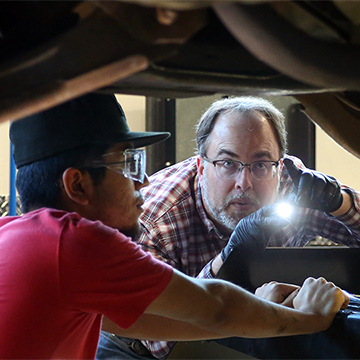 A JCCC student and his instructor use a flashlight to inspect the undercarriage of a vehicle