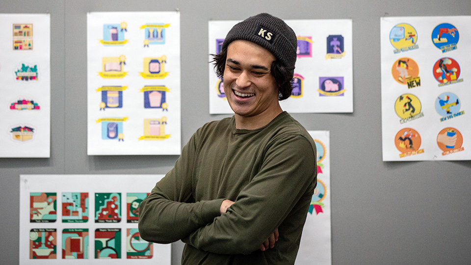 Smiling student standing in front of a display board of student graphic design work