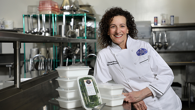 Diane Mora, wearing a white chef’s jacket that reads “Diane Mora, Kids Feeding Kids,” stands next to a metal culinary counter with her arm propped on food container boxed used in the Kids Feeding Kids program.