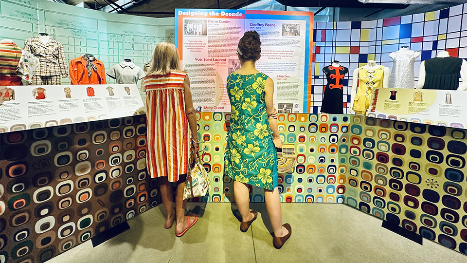 two women wearing 1960s apparel with their backs to the viewer look at an exhibition of historical 1960s apparel