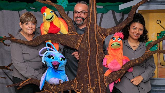 3 smiling puppeteers wearing microphones hold their colorful puppets up on a tree trunk