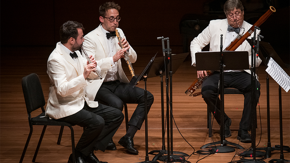 Three men wearing tuxedos sitting in chairs and playing instruments on a stage