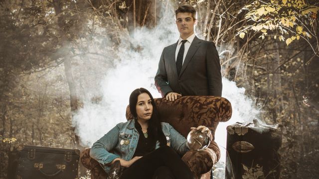 A woman sits in a chair holding a crystal ball. A man in a suit stands behind her. Both are looking at the camera. There is fog behind them and the setting is the woods.