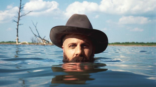 Marty Bush rising up out of a lake while wearing a cowboy hat. He is submerged up to his beard and facing the viewer.